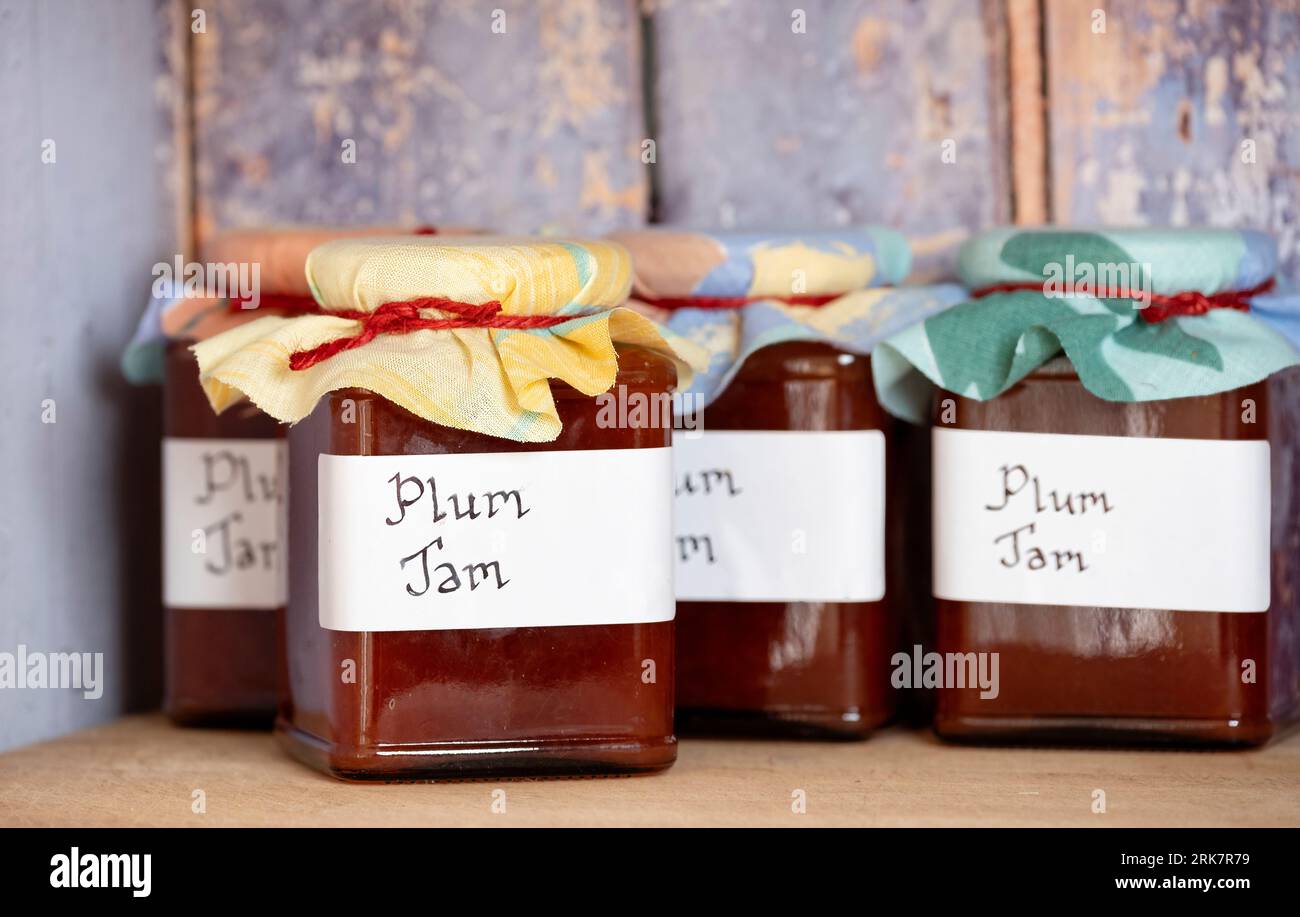 Freshly made jars of  homemade plum jam, with handwritten labels and cloth toppers placed on a shelf in a rustic wooden store cupboard Stock Photo