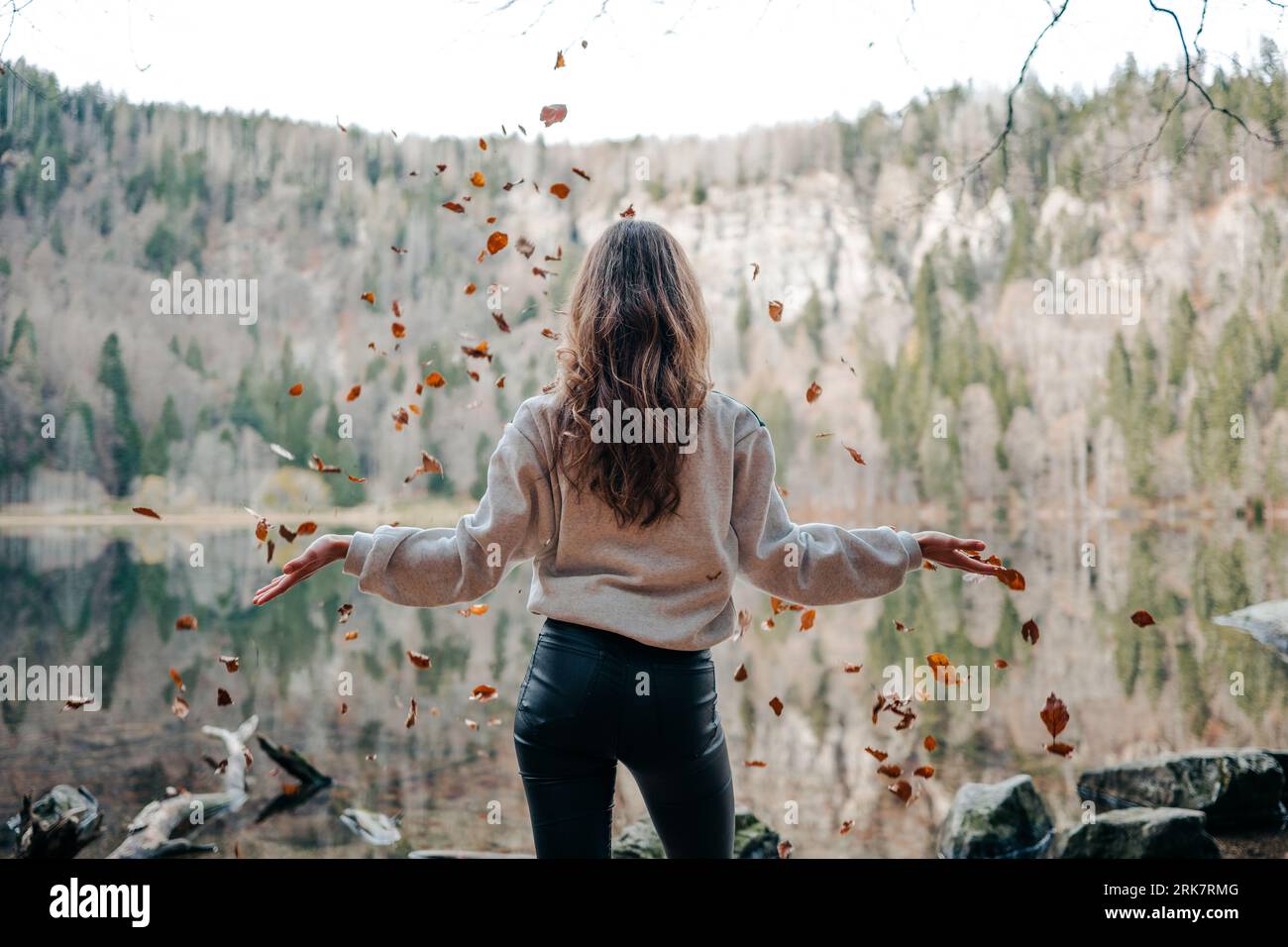 A young woman wearing tight jeans and a sweater, standing in front of a lake, throwing leaves. Stock Photo
