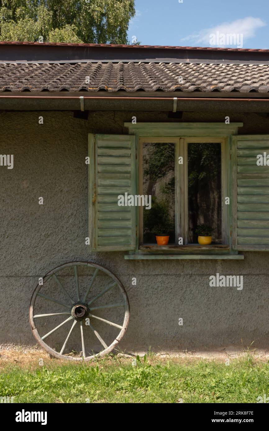 Picturesque garden shed or small cottage with old variate wheel leaning against the wall and window shutters open Stock Photo