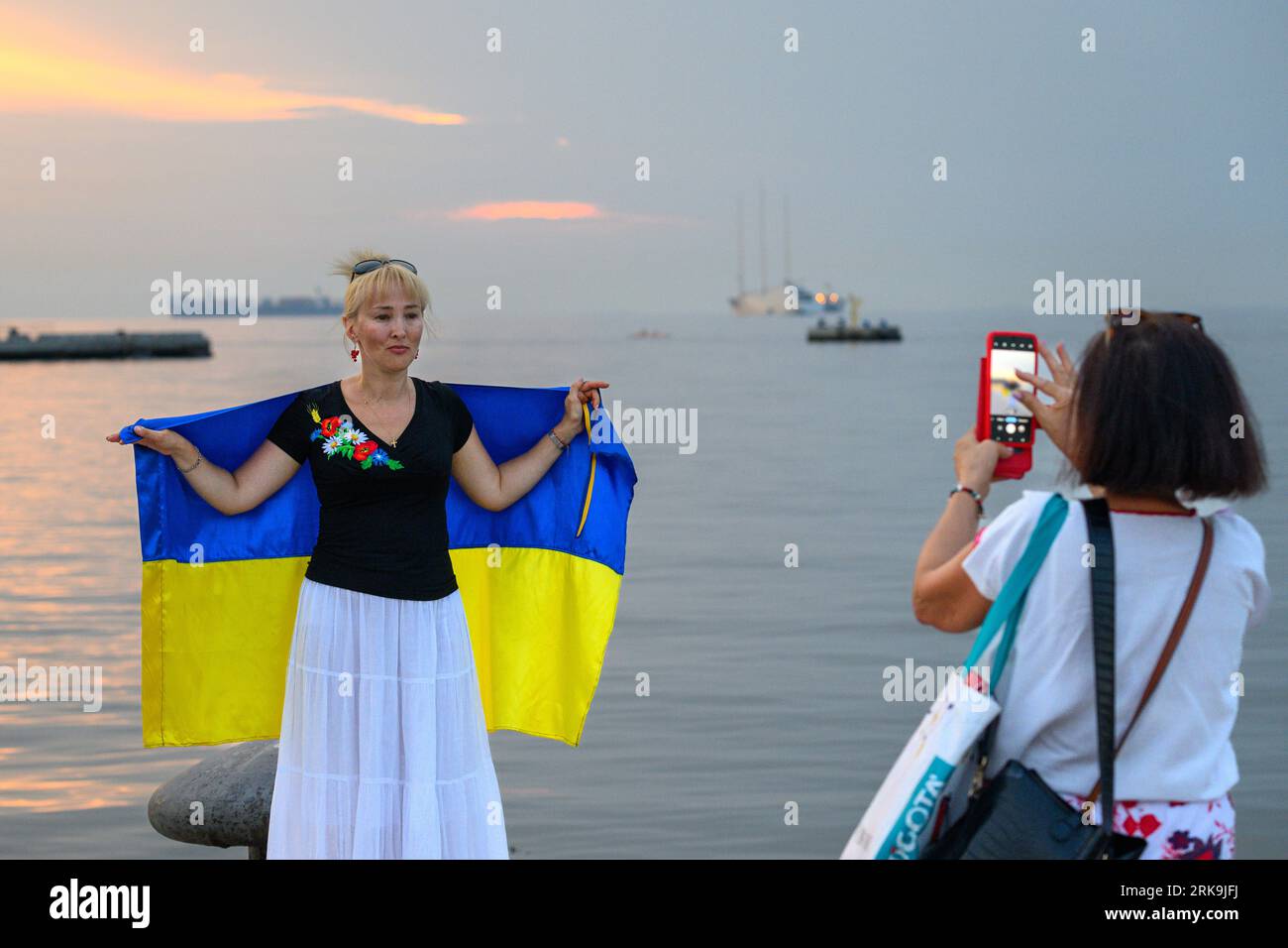 Trieste, Italy, 24 August 2023. Ukrainian women display their national flag to mark Ukraine's 32nd Independence Day in the Adriatic Sea port of Trieste, Italy. Ukrainian residents held a demonstration in the central Piazza Unità d'Italia was part of a 'Chain of Unity' organized by Ukrainian communities worldwide to show their resilience, dignity and support for their country, invaded by Russia exactly 18 months ago. Far away in the horizon is the largest megayacht in the world, the 530 million euro Sailing Yacht A belonging to Russian oligarch Andrey Igorevich Melnichenko, seized in March 20 Stock Photo