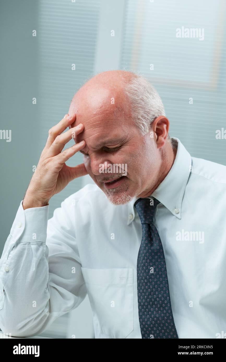 Senior man in business attire holds head, closes eyes, and clenches teeth. Evident pain or regret of failure. Emotions of frustration, disappointment, Stock Photo