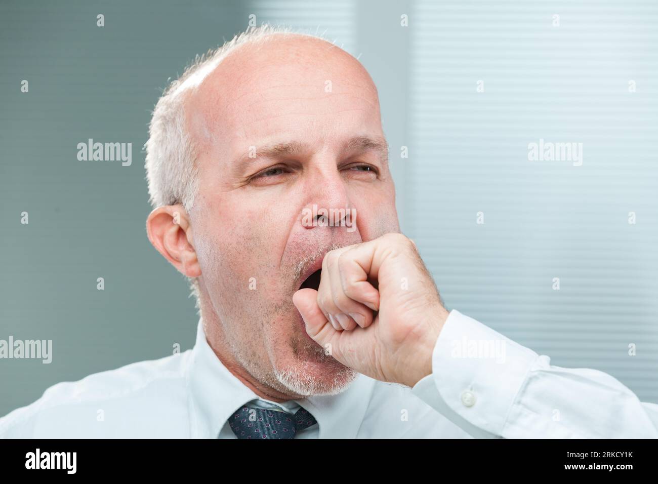 Aged clerk yawns, covering his mouth ineffectively. The close-up reveals a man tired, perhaps bored, needing oxygen or hinting at a transition. Dresse Stock Photo