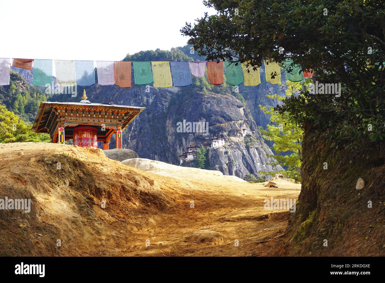 A prayer wheel pagoda to one side and colorful prayer flags above the trail frame a view of Tiger's Nest Monastery perched on the mountainside ahead Stock Photo