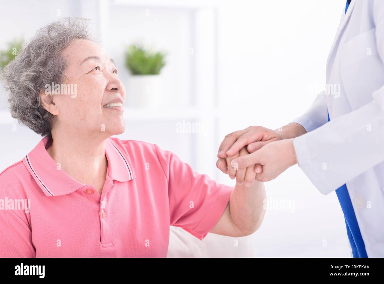 physician leaning forward to smiling elderly lady patient holding her hand in palms. Woman caretaker in white coat supporting encouraging old person Stock Photo