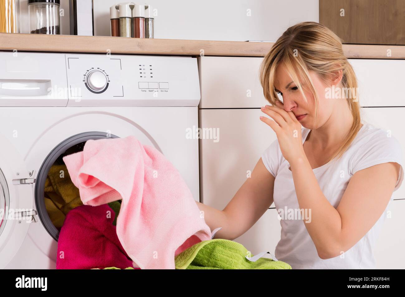 Young Woman Looking At Smelly Clothes Out Of Washing Machine In Kitchen