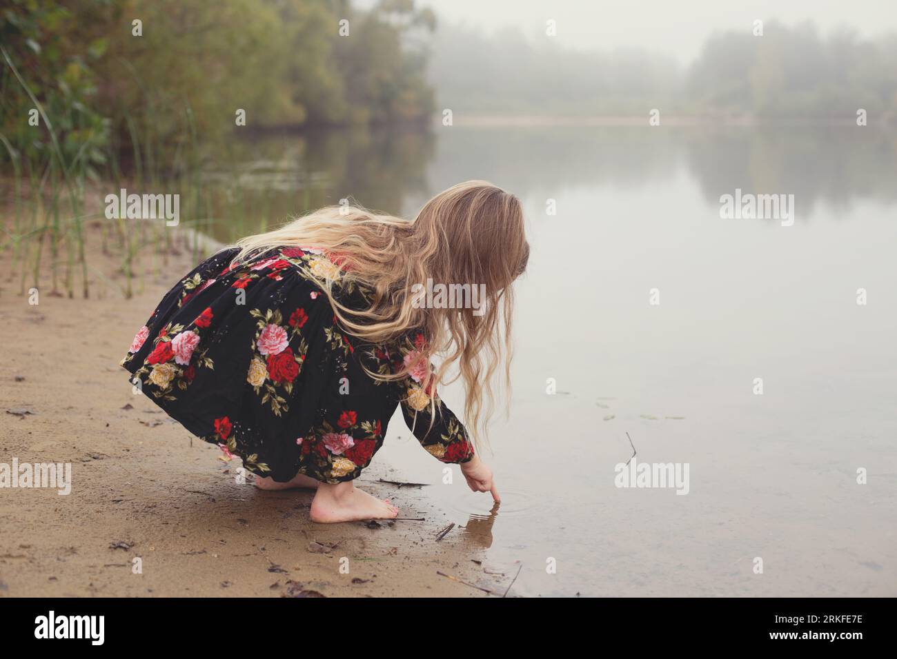 Little girl on a lonesome and foggy adventure dips finger in pond Stock Photo