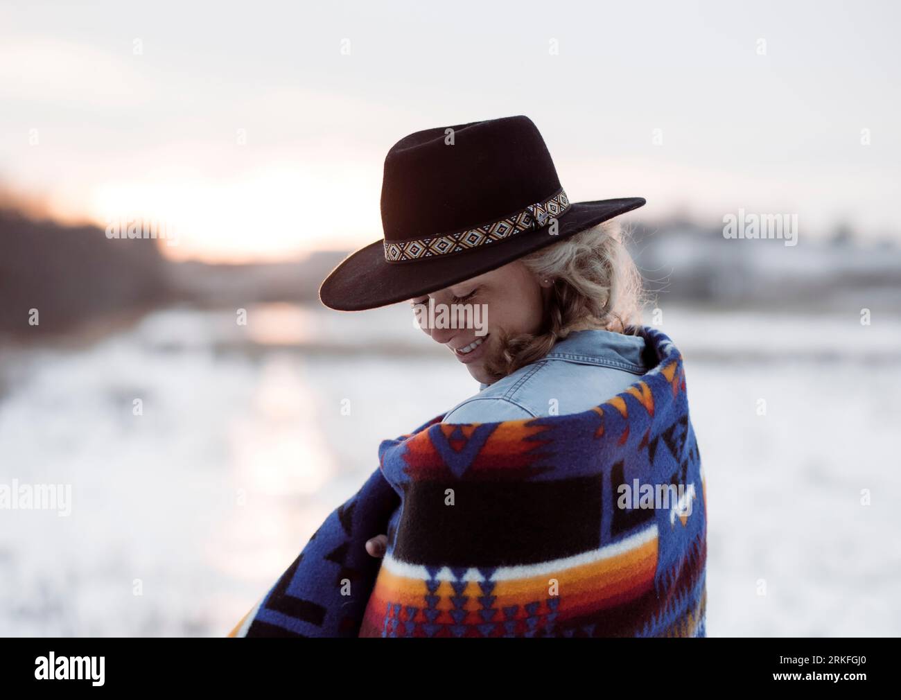 woman in a hat wrapped in a blanket looking down smiling at sunset Stock Photo