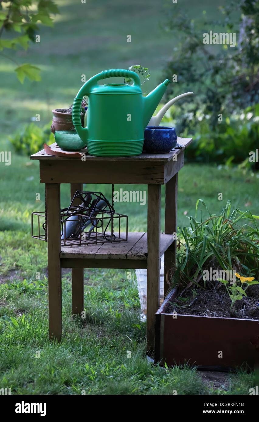 Wood table with watering can, pots and gardening tools still life next to a flower garden on a summer morning in Taylors Falls, MN Stock Photo