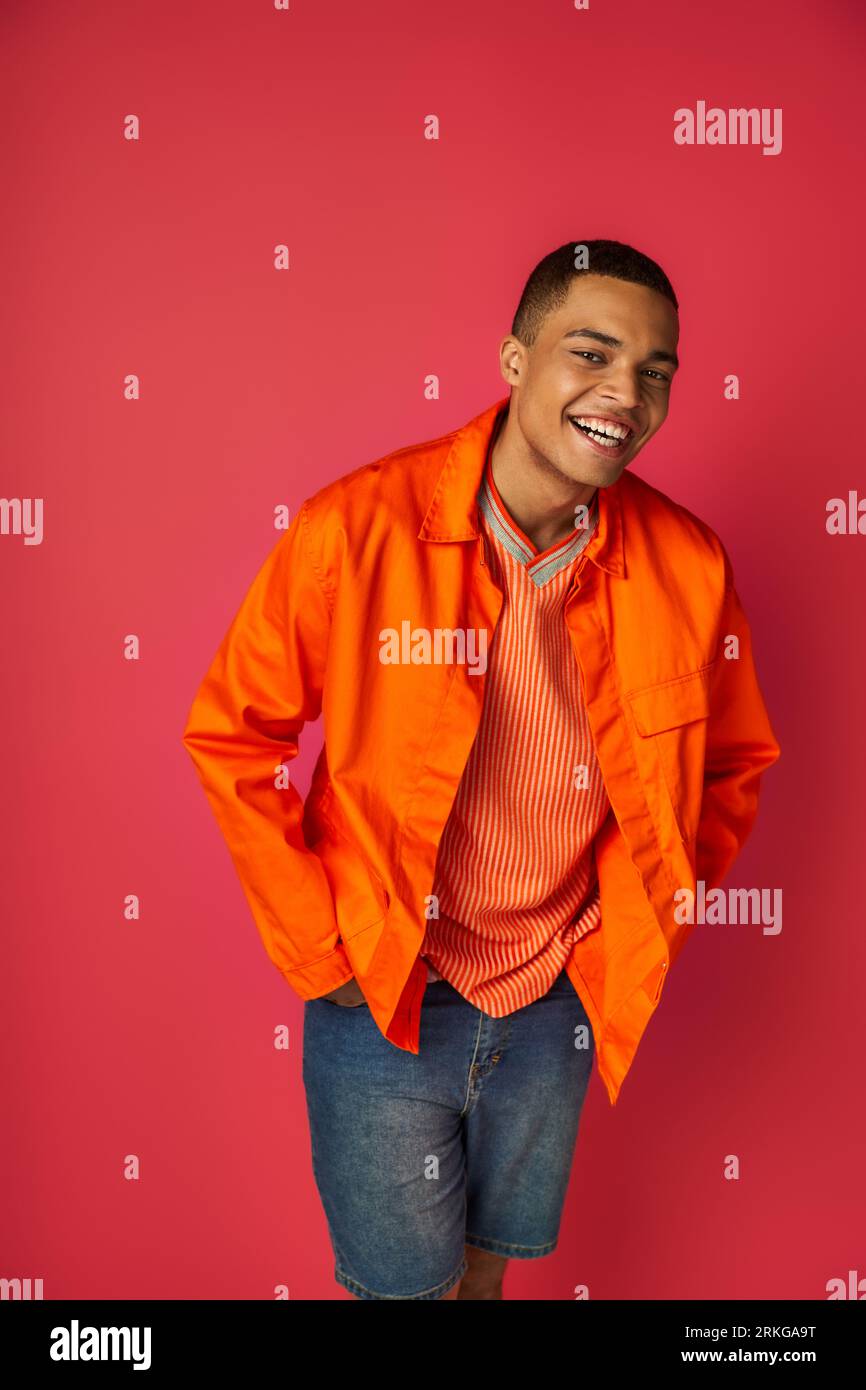 joyful african american guy in orange shirt, with hands in pockets