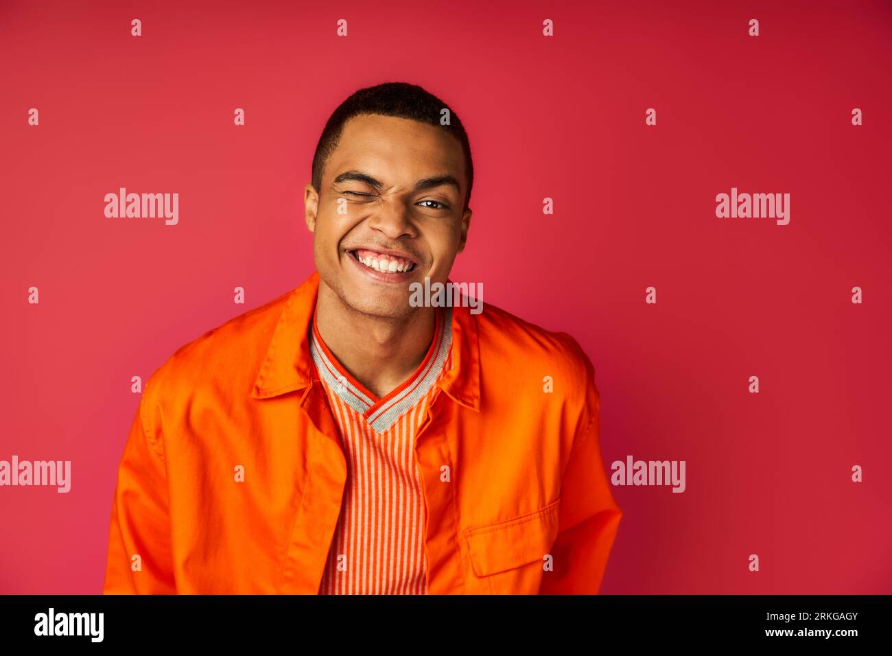 cheerful african american guy in orange shirt winking at camera on red