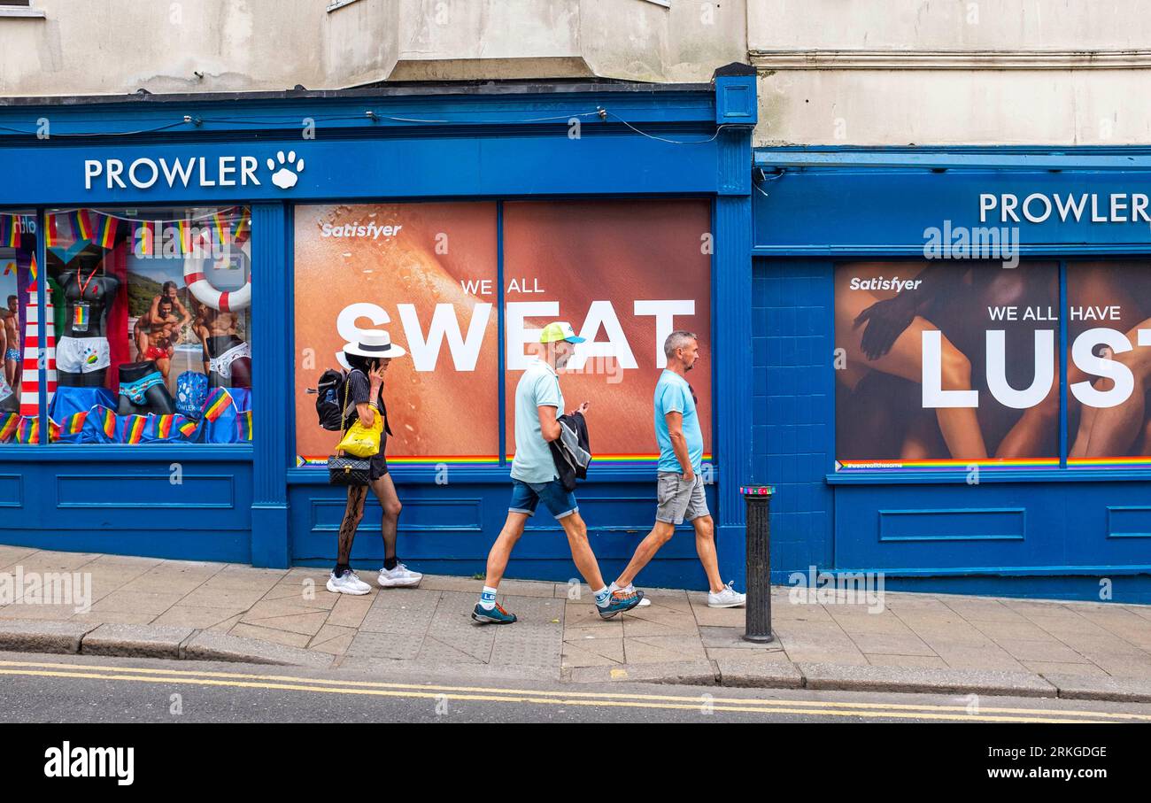 People walking past the Prowler specialist gay shop in St James's Street Kemptown Brighton which has the slogans We All Sweat and We All Have Lust on the windows , Sussex UK Stock Photo