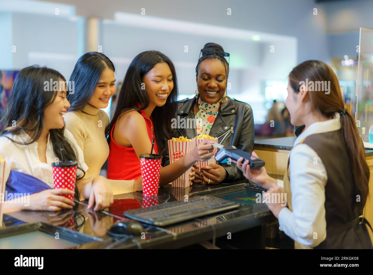 Effortlessly, interracial female friends pay for movie tickets using a credit card at the counter, embracing contactless convenience for a seamless ci Stock Photo