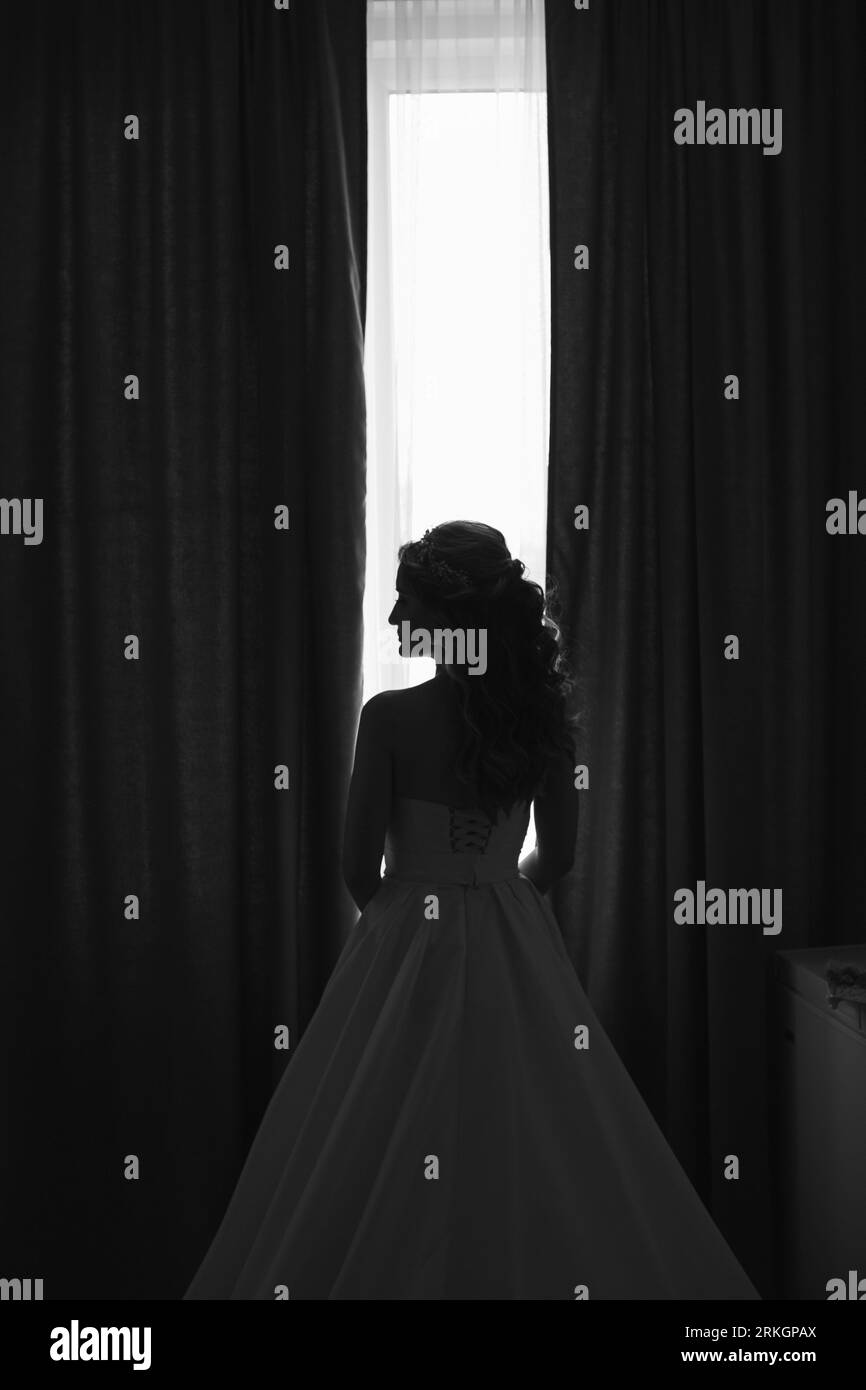 A full-length shot of a bride standing in profile, admiring the view from a tall window in her wedding dress Stock Photo