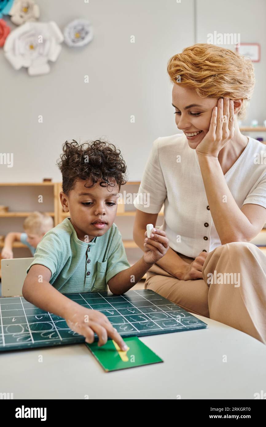 happy teacher observing african boy pointing at number near chalkboard