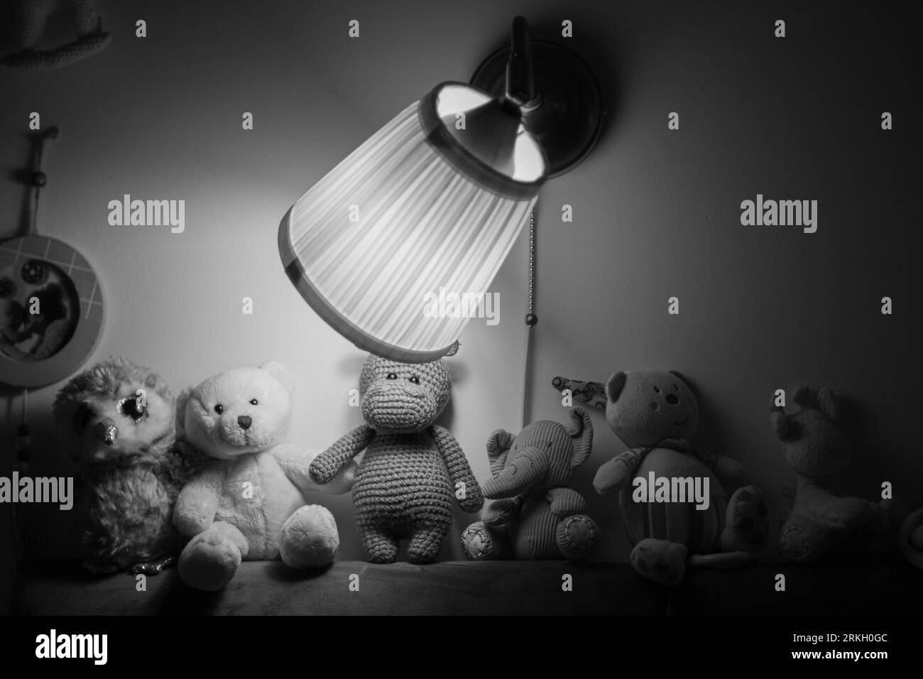 An arrangement of stuffed animals gathered around a desk illuminated by a warm light in grayscale Stock Photo