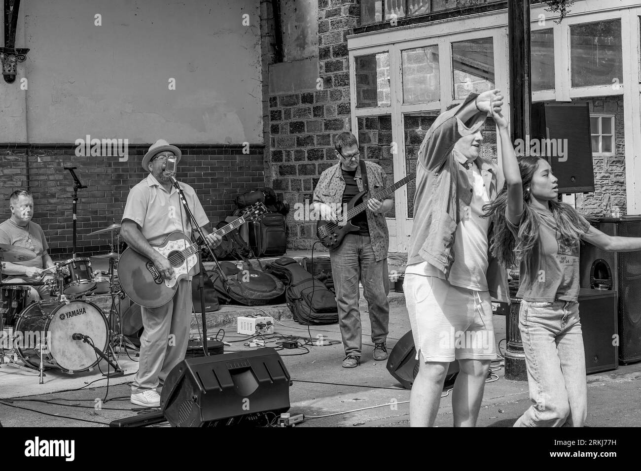 A couple dances in front of a band, which consists of a lead singer/guitarist, bass player, and drummer, in Crescent Gardens, Harrogate, UK. Stock Photo