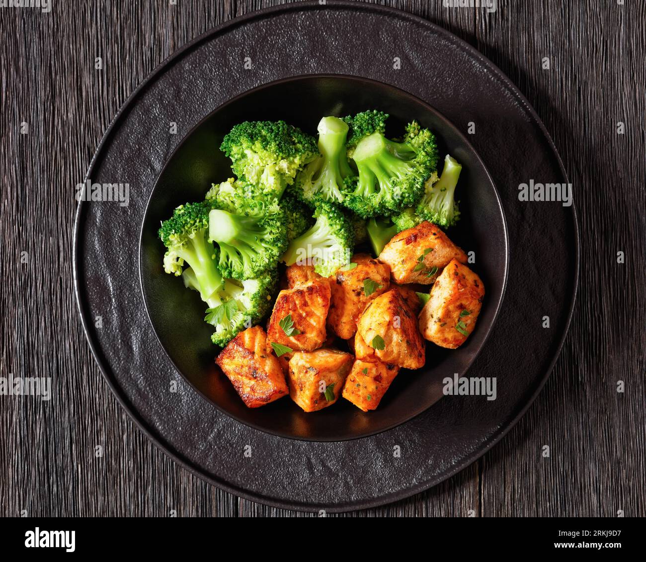 baked salmon bites with steamed broccoli florets in black bowl on dark wooden table with sriracha mayo sauce, horizontal view from above, close-up Stock Photo