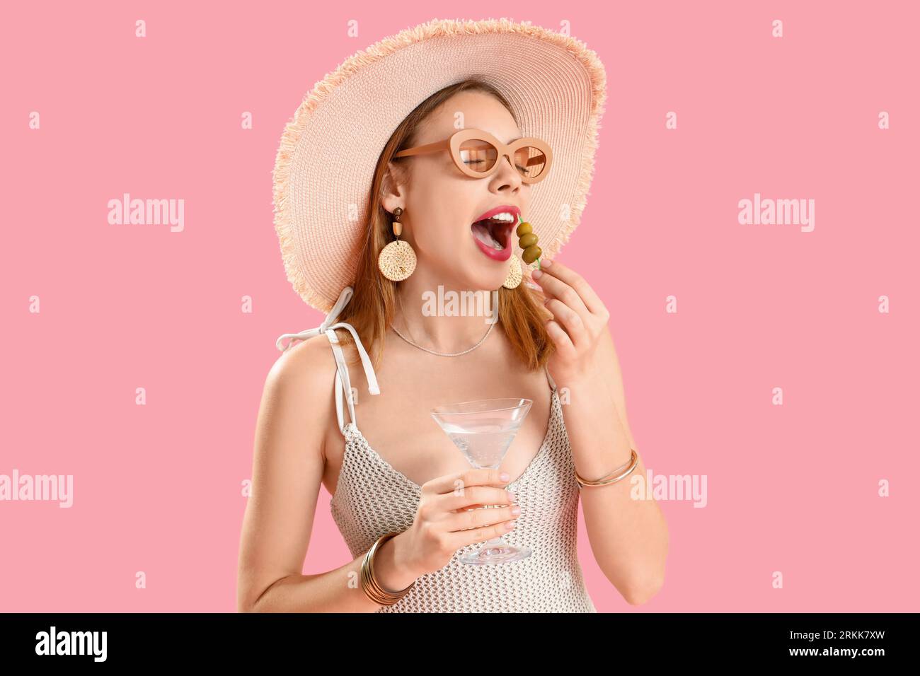 Beautiful young woman in wicker hat with glass of martini eating olives on pink background Stock Photo