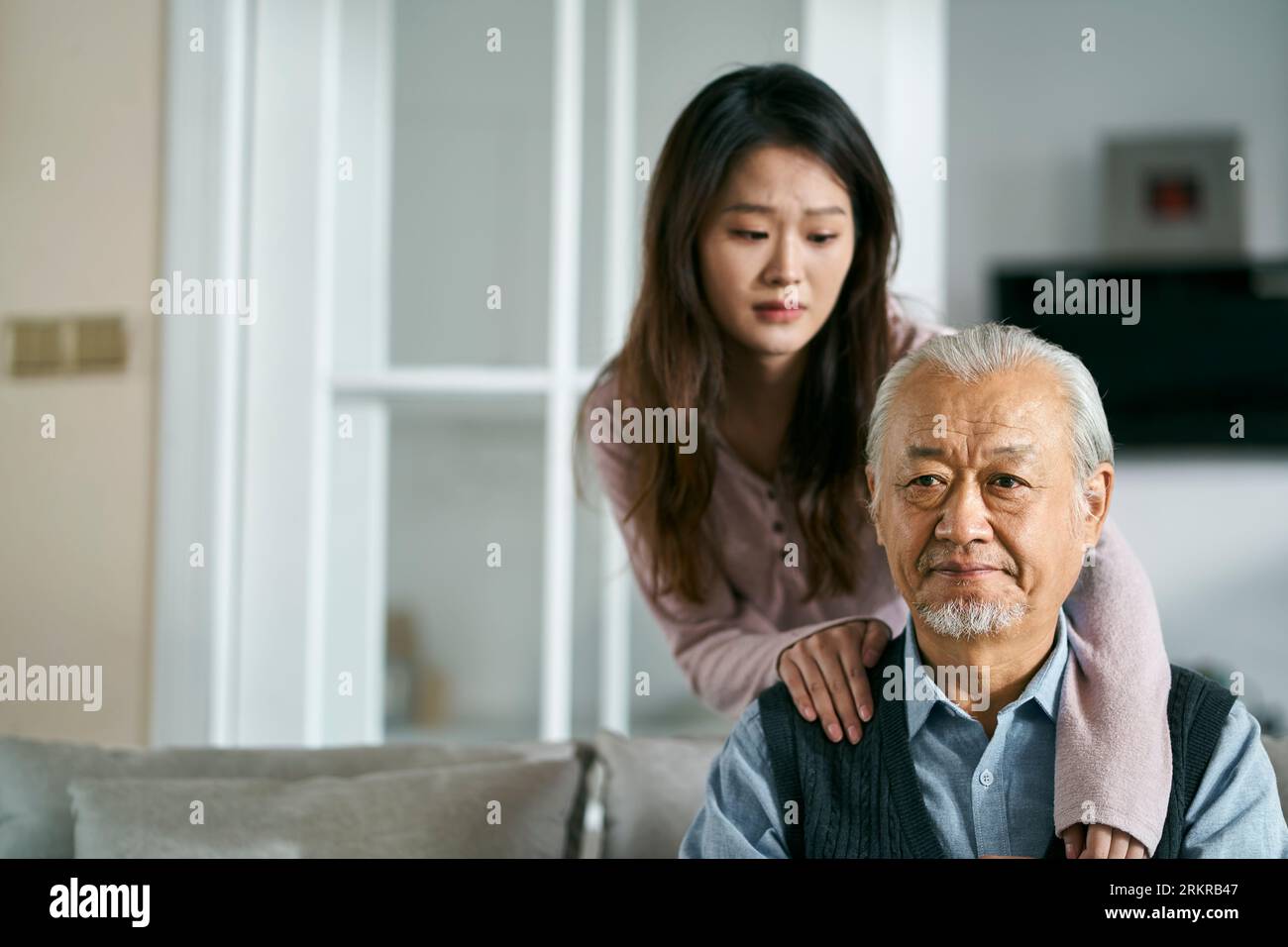 sad senior asian father sitting on couch in living room at home consoled by adult daughter Stock Photo