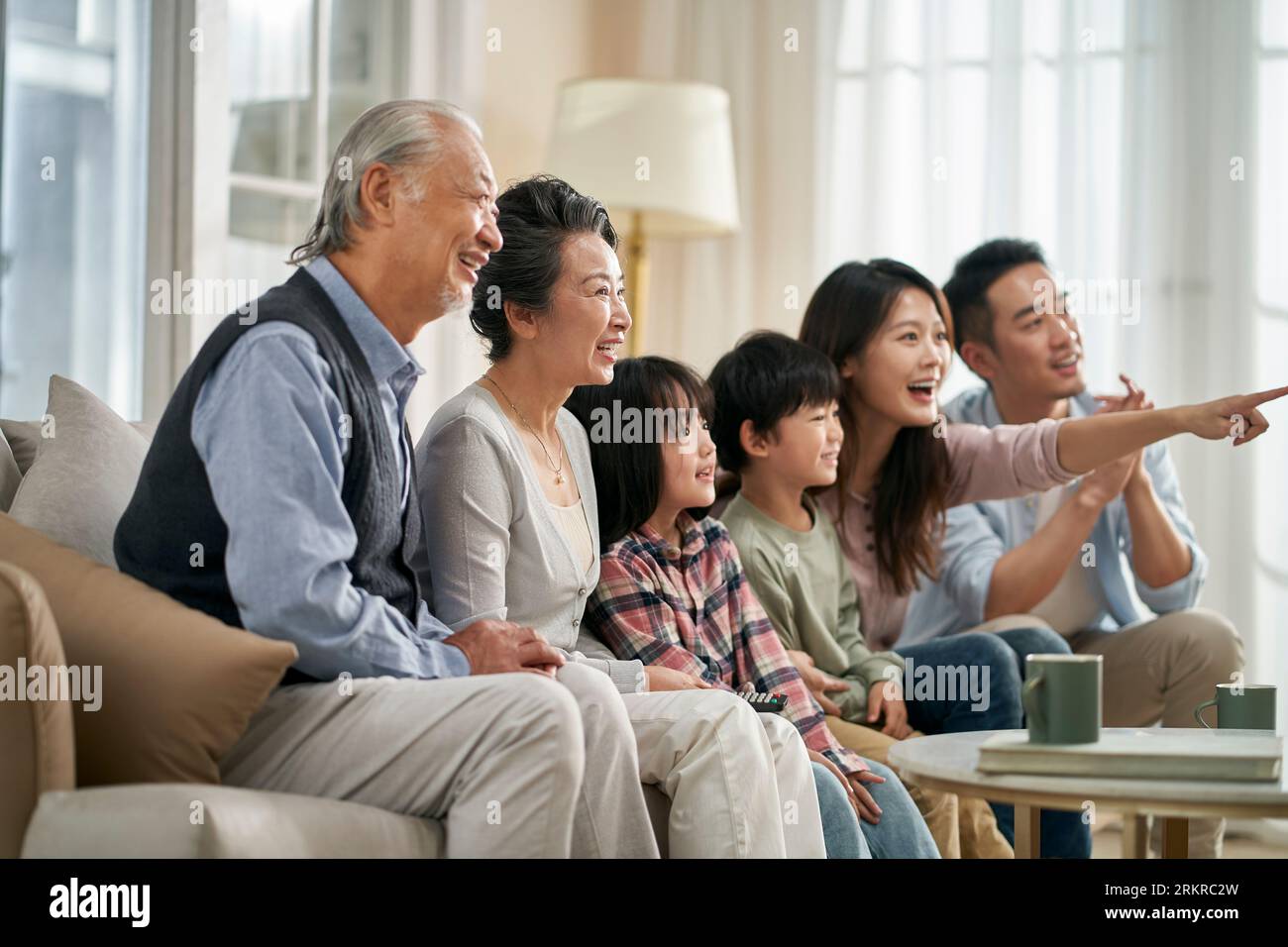 three generation asian family sitting on couch at home watching tv together happy and smiling Stock Photo