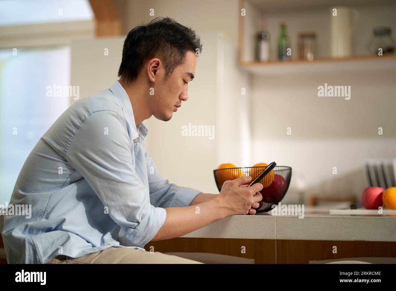 young asian adult man sitting at kitchen counter using mobile phone Stock Photo