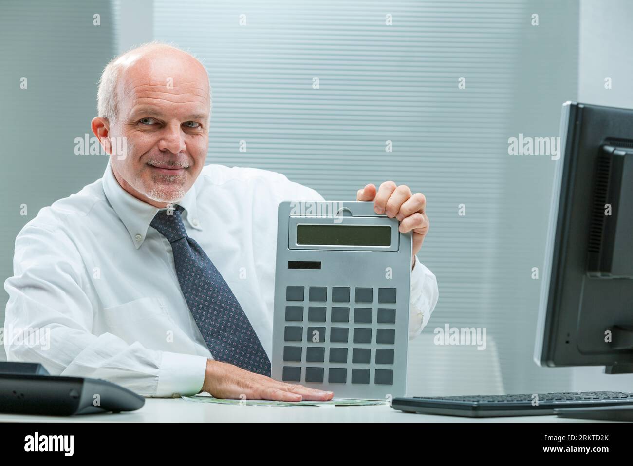 senior businessman in white shirt and tie showcases a cunning glance, clutching a sizable calculator; suggests misleading through manipulated data Stock Photo