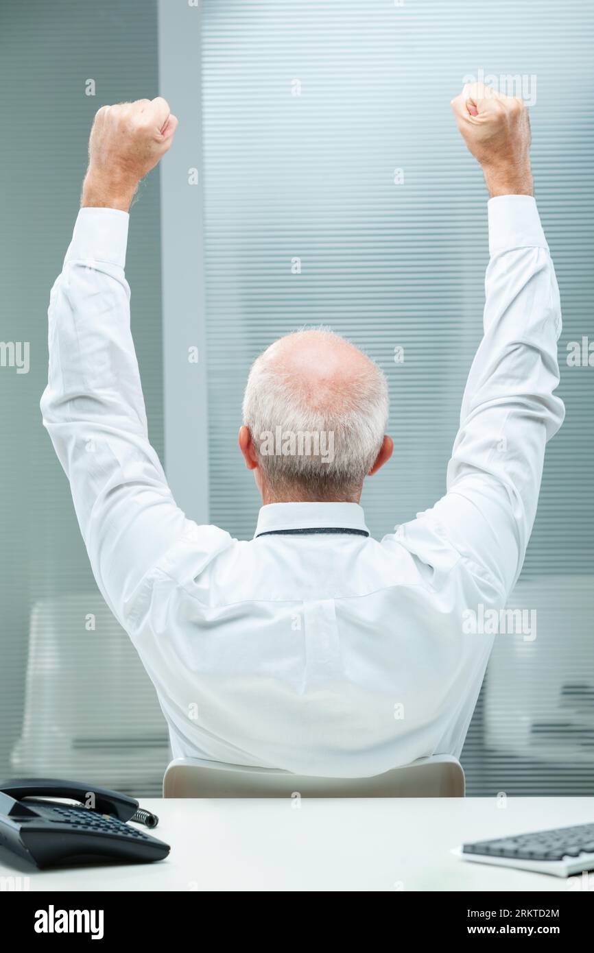 senior businessman, viewed from behind, raises his arms in sheer joy and triumph. The visible hairless center is framed by white hair, symbolic of age Stock Photo