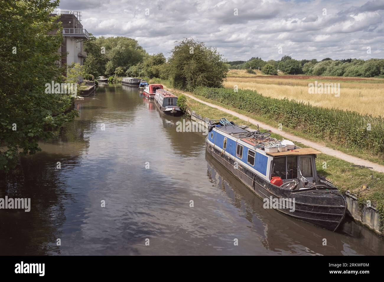 six narrowboats line one side of the river stort near Harlow England among beautiful trees and farmland on a sunny day with a cloudy sky background Stock Photo