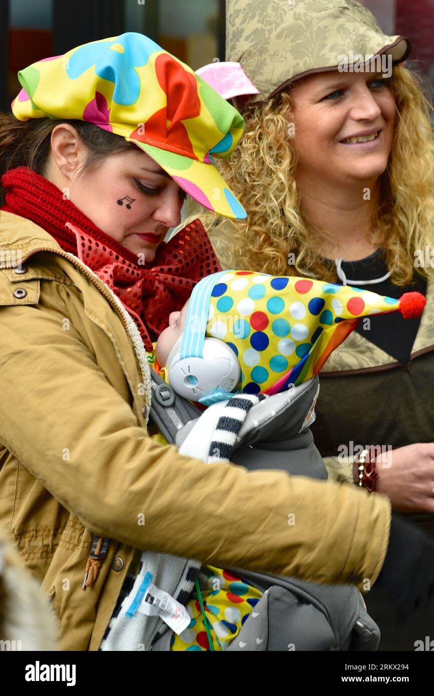 Mother with a colorful carnival cap and a music note on her cheek looks at her baby in a carrier with a somewhat concerned look. Stock Photo