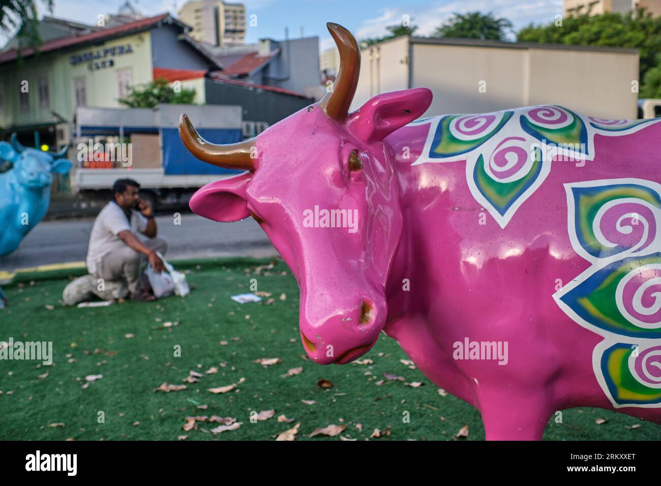 A worker from India sits near a colorful cow statue in a small park. in Little India area, Singapore, the 'grass' actually being artificial turf Stock Photo