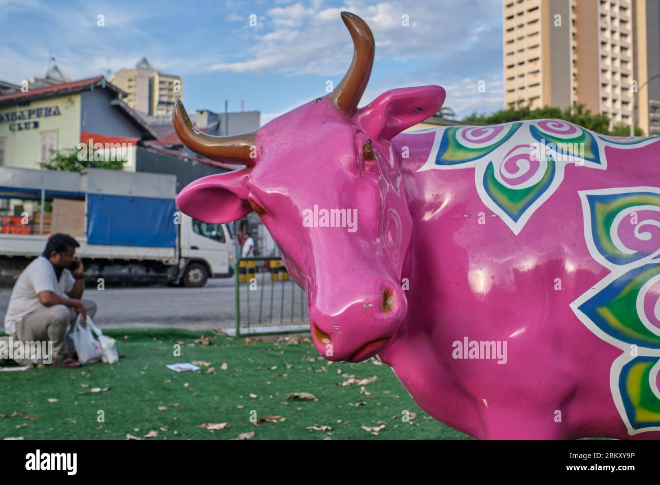 A worker from India sits near a colorful cow statue in a small park. in Little India area, Singapore, the 'grass' actually being artificial turf Stock Photo
