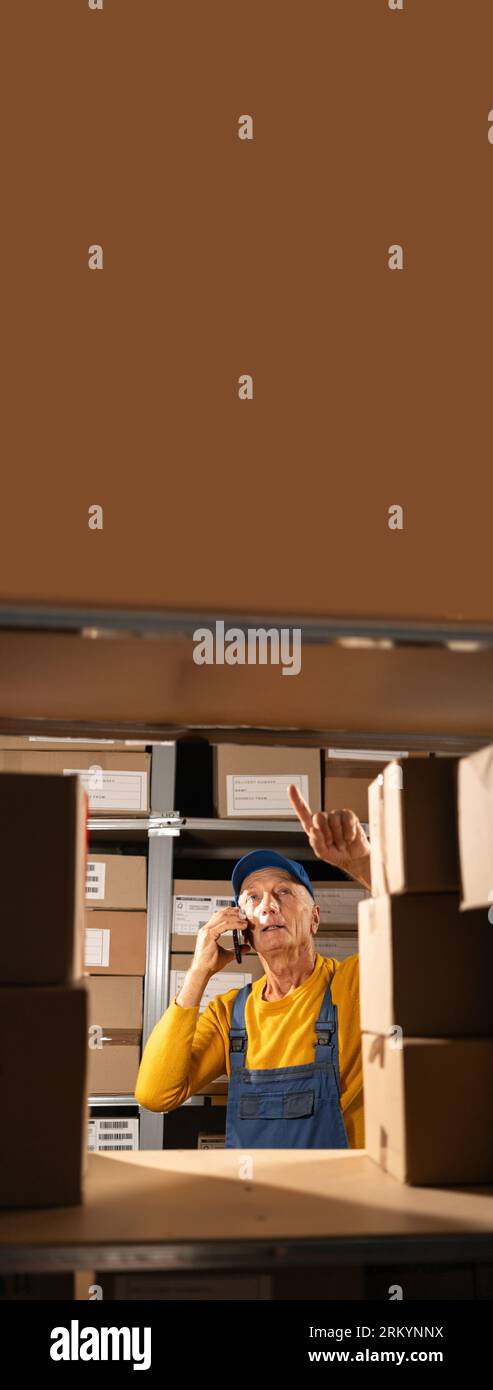 Warehouse elderly worker counting shipment on shelves align with total box number in stock on smartphone to confirm the inventory before delivery. Stock Photo