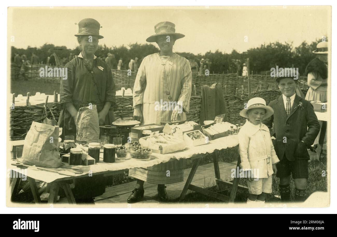 Original early 1900's era postcard of summertime village fete, with a bring and buy stall. A lady committee member stands besides her stall with a stallholder, the stall displays sells jams, fruit nougats, damsons by the pound - weighing scales in background. Two young customers stand next to the trestle table laid out with wares for sale. The young boy wears an HMS Defender sailors hat, and the other child, a flat cap. Circa 1919 /1920, U.K. Stock Photo