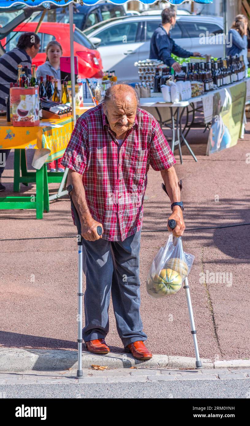 Elderly man holding bag of melons crossing road using medical walking canes - Preuilly-sur-Claise, Indre-et-Loire (37), France. Stock Photo