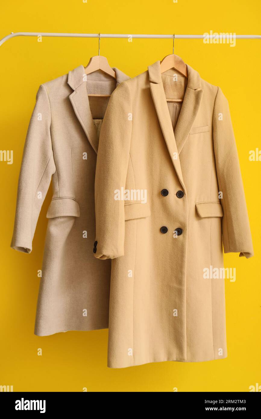 Stylish coats hanging on rack against yellow background, closeup Stock Photo