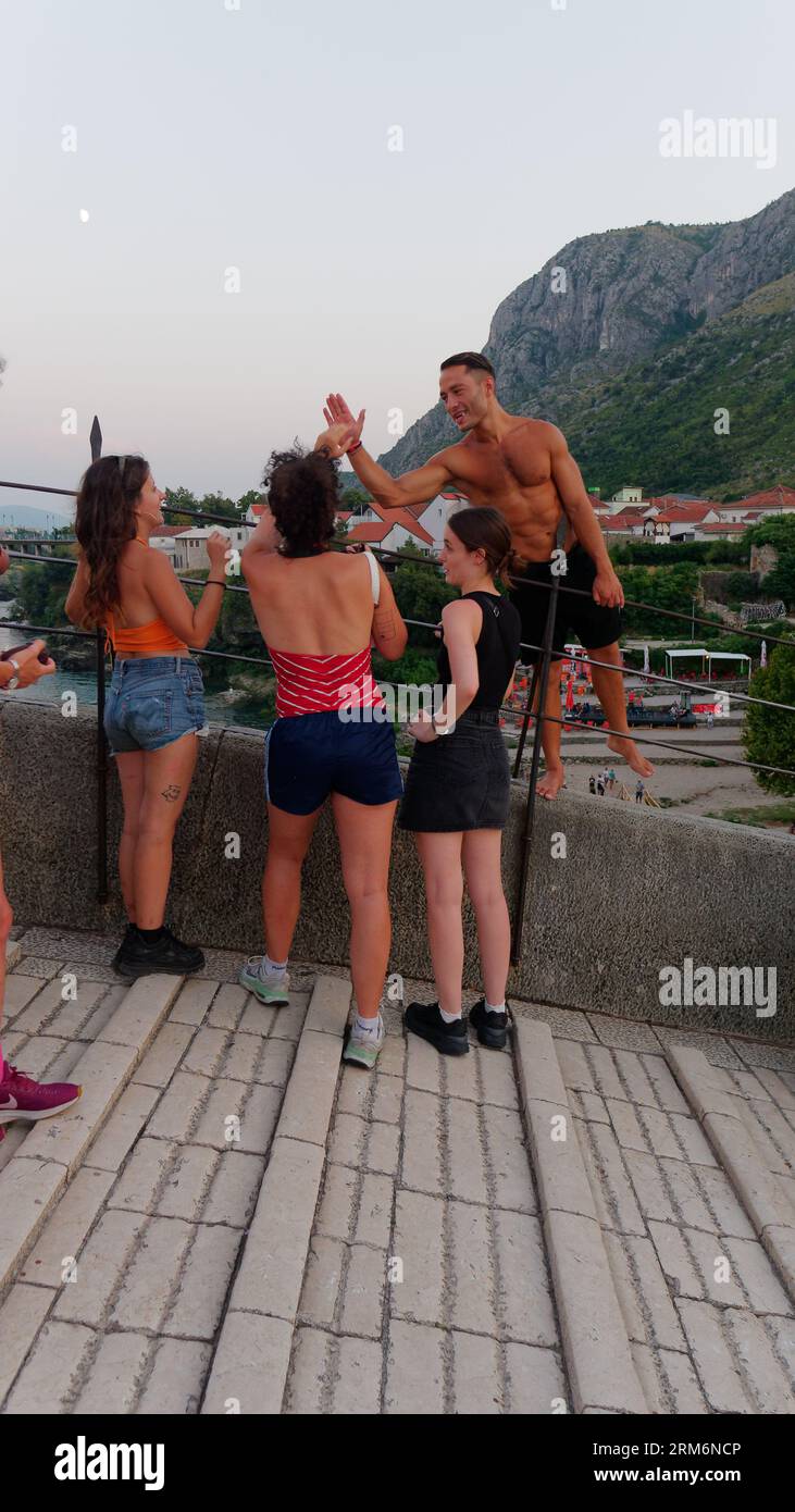 Muscular local man high fives tourist females on Stari Most (Old Bridge) with Neretva River below. Mostar, Bosnia and Herzegovina, August 22, 2023. Stock Photo