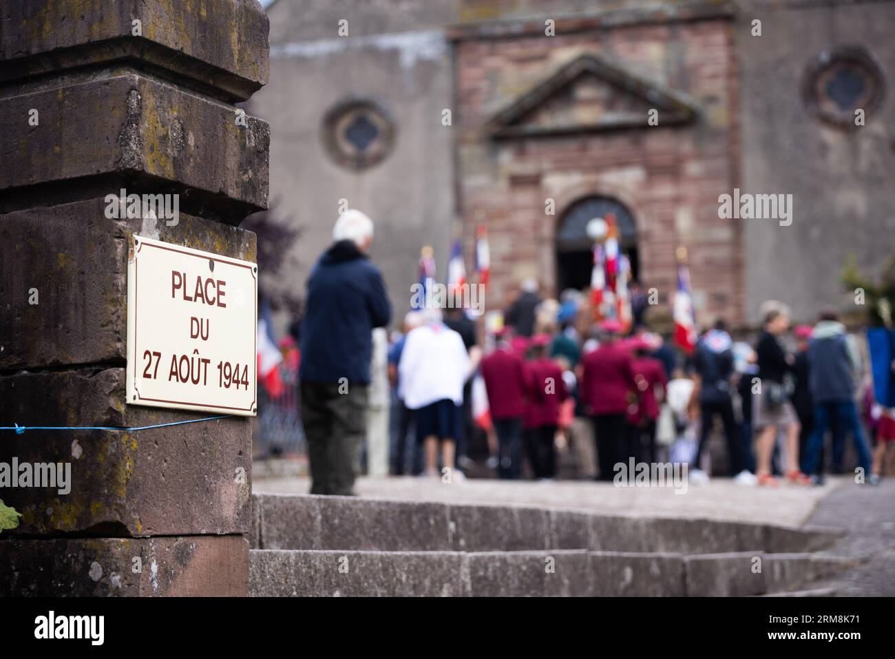 Pexonne, France. 27th Aug, 2023. A street sign marks the 'Place du 27 Aout 1944' (engl.: 'Square of August 27, 1944') while in the background people gathered for a memorial service. In late August 1944, German soldiers invaded the French village of Pexonne and abducted dozens of villagers. The action was led by SS Hauptsturmführer Erich Otto Wenger. Now Wenger's grandchildren came to town for the first time to take part in a memorial service for the victims of the crime. Credit: Philipp von Ditfurth/dpa/Alamy Live News Stock Photo