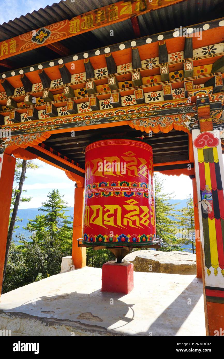 An oversized prayer wheel sheltered by a pagoda roof with typical Bhutanese painted architecture with trees and sky visible beyond near Paro, Bhutan Stock Photo