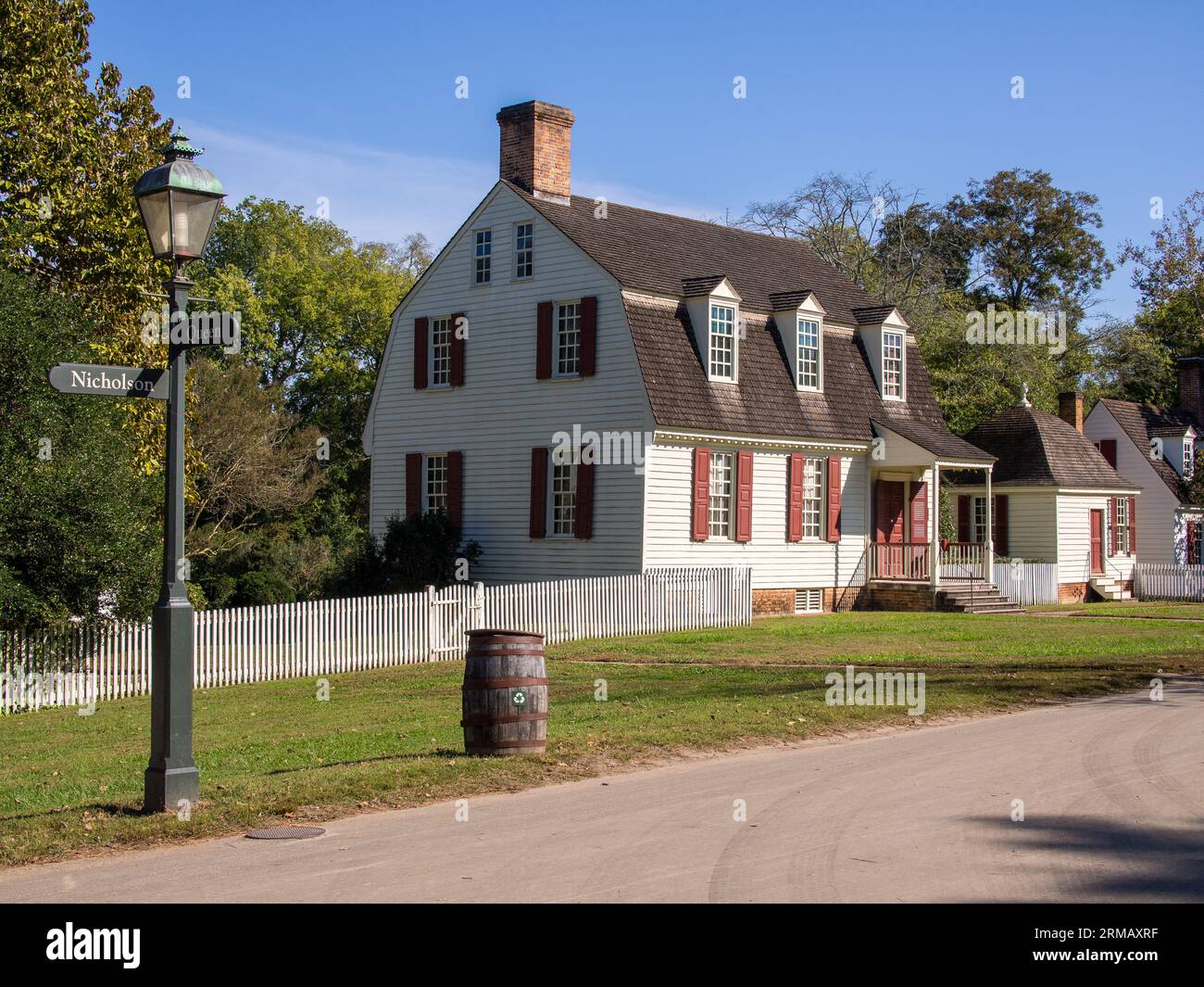 Elegant white colonial home, dual-story design, wooden barrel, charming white fence, street signs, vintage lamp post, set against a vivid blue sky in Stock Photo