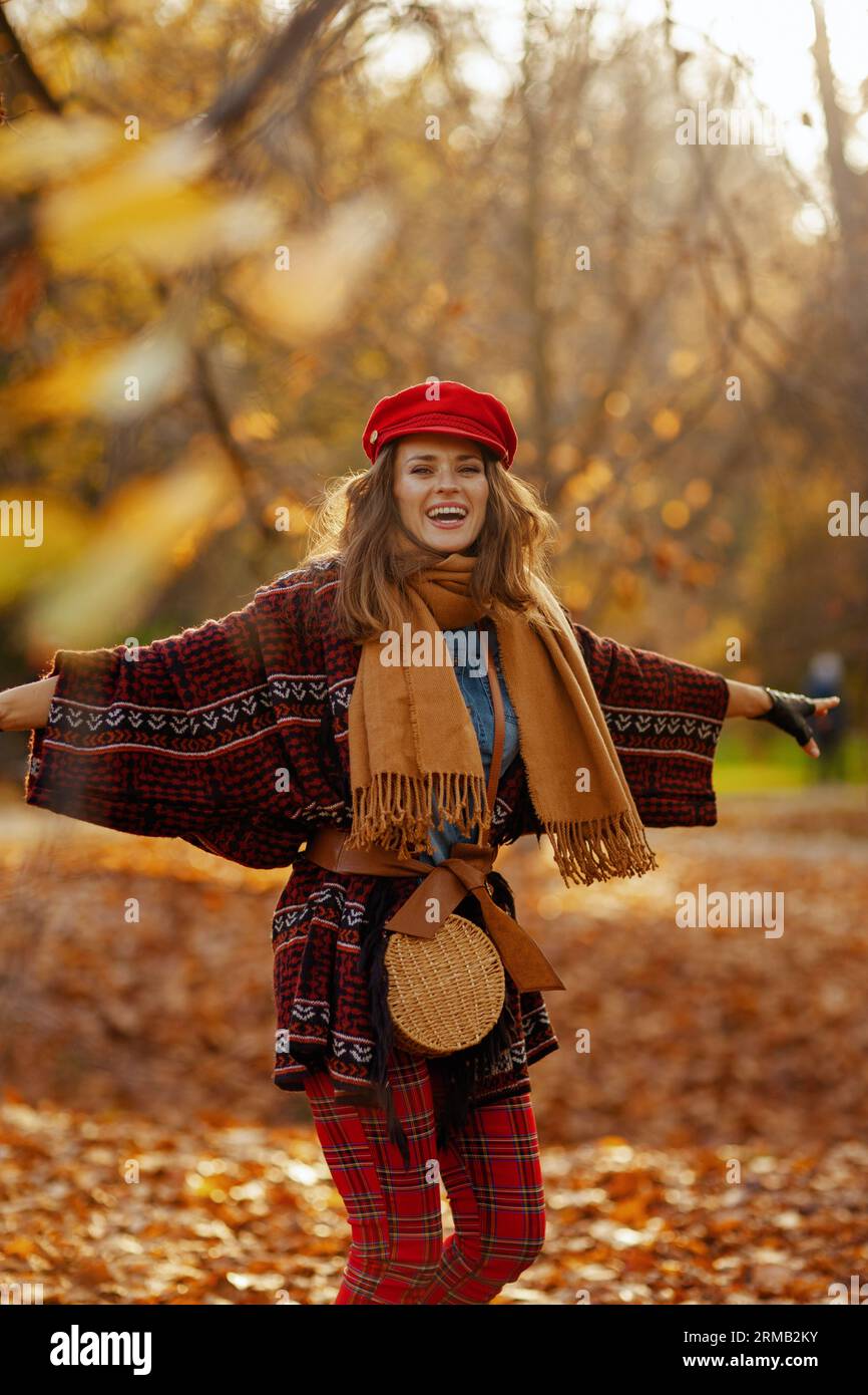 Hello autumn. smiling trendy 40 years old woman in red hat with scarf, gloves and bag rejoicing in the city park. Stock Photo