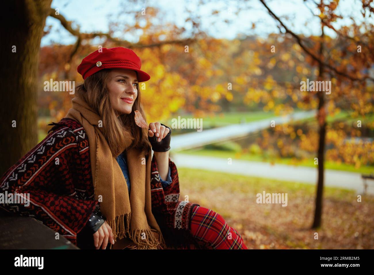 Hello autumn. pensive modern woman in red hat with autumn leaf and scarf sitting on bench in the city park. Stock Photo