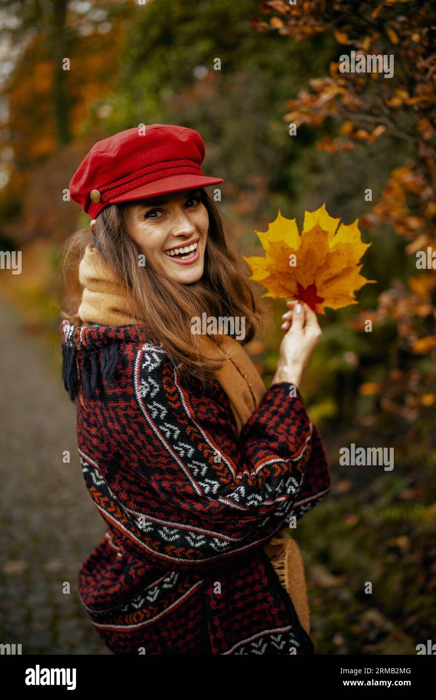 Hello autumn. Portrait of smiling elegant middle aged woman in red hat with autumn leafs and scarf walking in the city park. Stock Photo