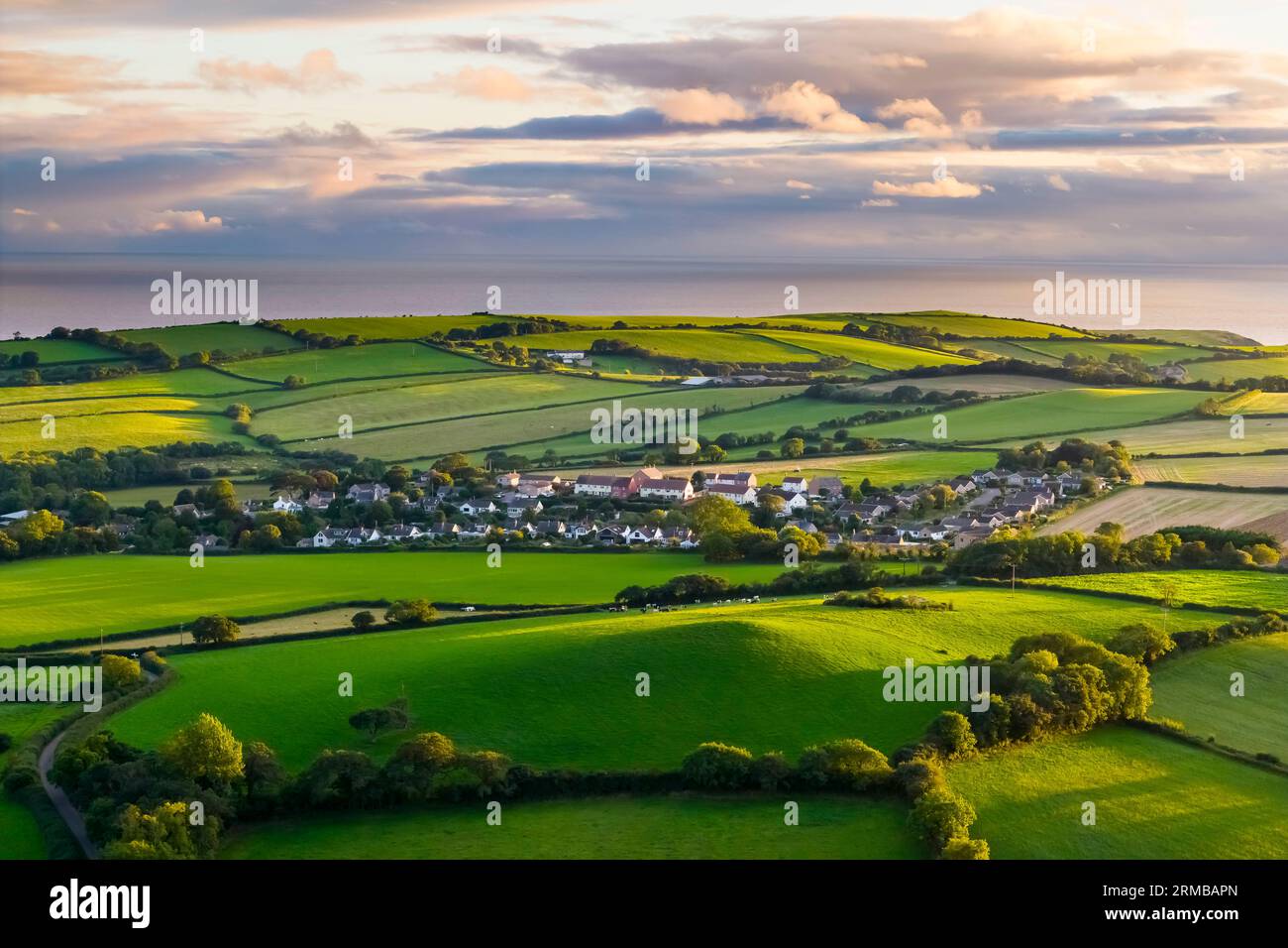 Shipton Gorge, Dorset, UK.  27th August 2023.  UK Weather.  Aerial view across the fields looking towards the village of Shipton Gorge in Dorset as golden hour sunlight bathes the countryside in sunshine shortly before sunset with dark clouds in the distance. Picture Credit: Graham Hunt/Alamy Live News Stock Photo