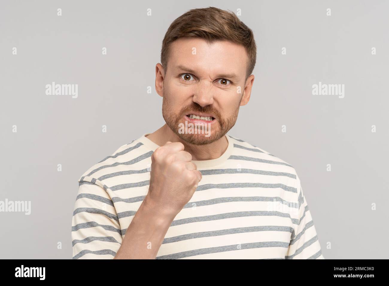 Angry aggressive middle aged man clenched fist has evil face looking at camera on grey background. Stock Photo