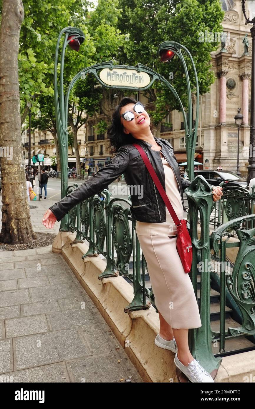 A happy stylish woman in a black leather jacket swings on the ornate cast iron art nouveau metro entrance at Fontaine Saint-Michel, Paris, France Stock Photo