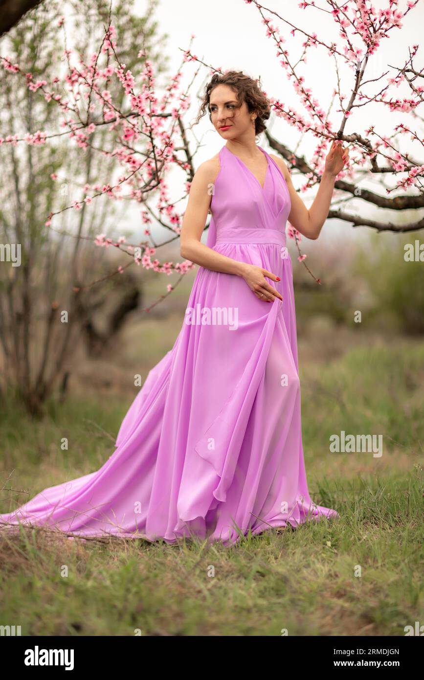 Woman peach blossom. Happy curly woman in pink dress walking in the garden of blossoming peach trees in spring Stock Photo
