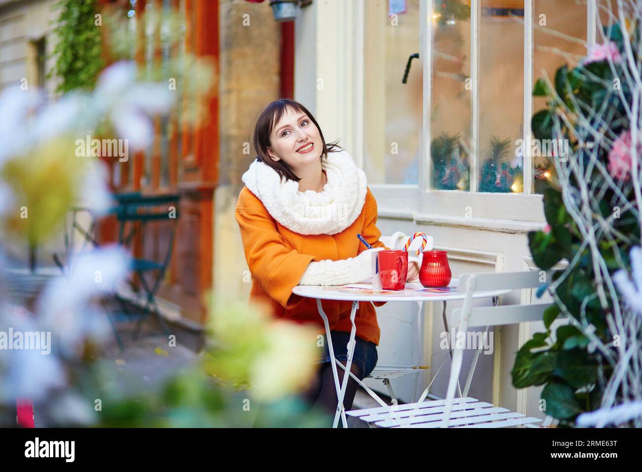 Happy young woman in Paris on a winter day, drinking coffee, tea or hot chocolate in Parisian outdoor cafe decorated for Christmas, writing postcards Stock Photo