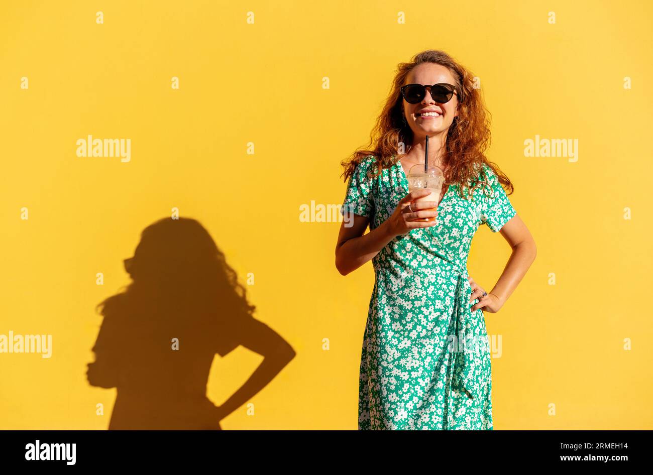 Young curly woman wearing trendy green dress and sunglasses holding glass of juice in hand and posing near yellow background. Stock Photo