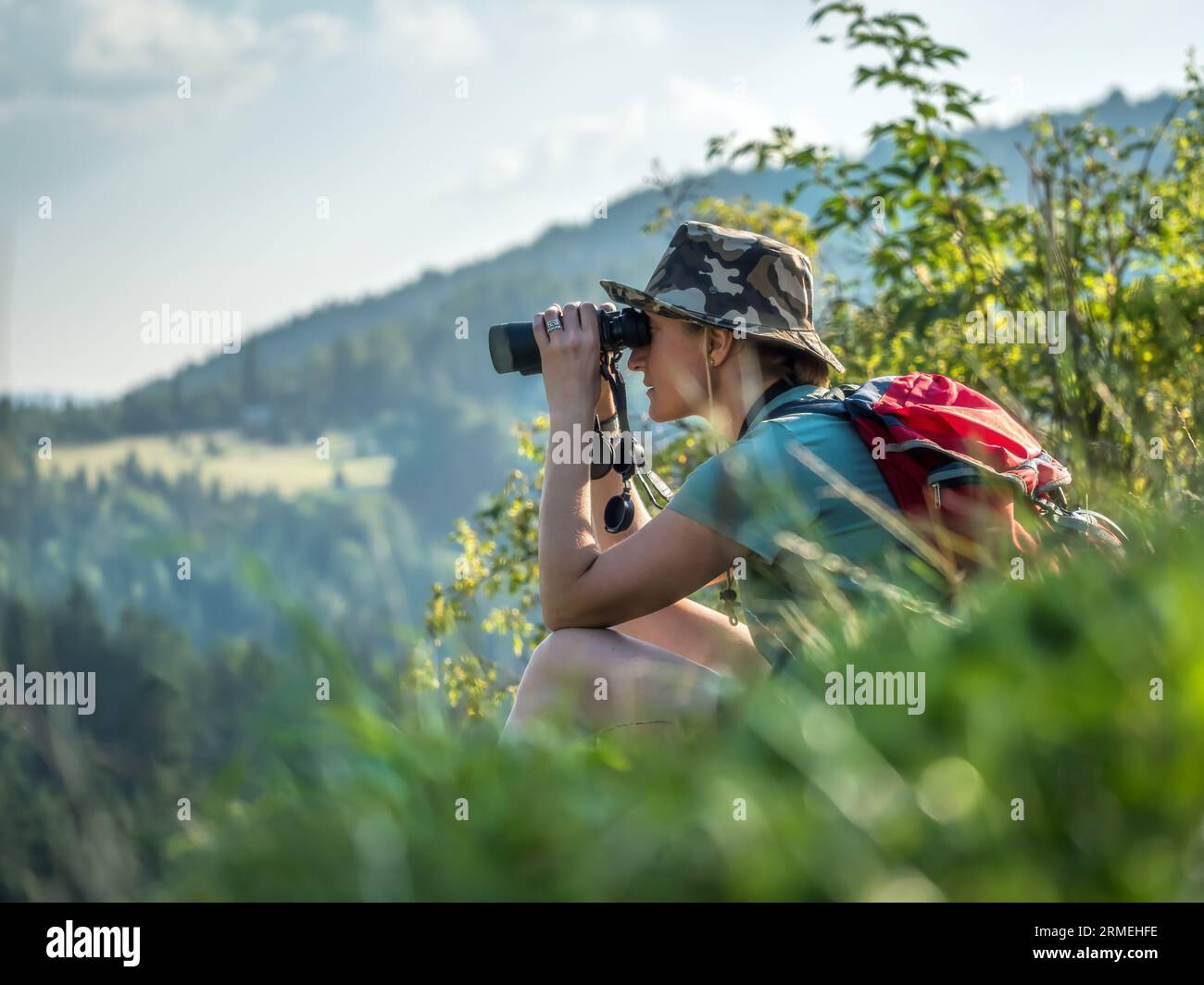Female tourist sitting on hillslide, admiring mountain vista through binoculars Stock Photo