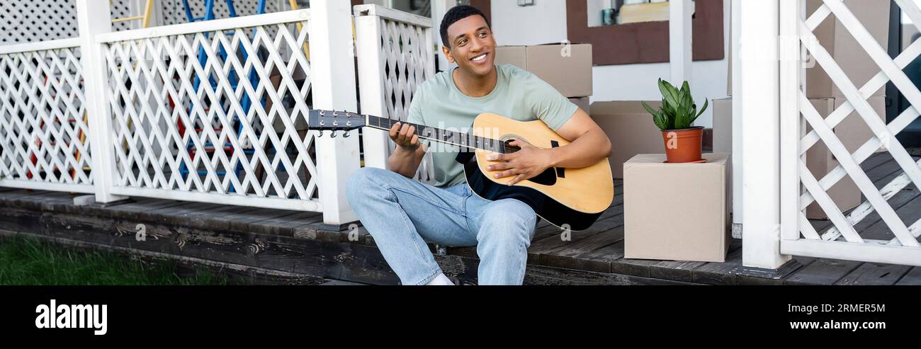 happy african american man playing acoustic guitar near carton boxes on  porch of new house, real estate, banner Stock Photo - Alamy